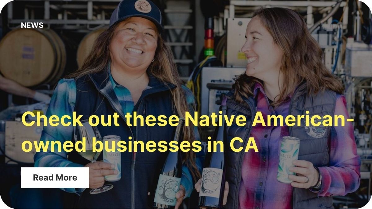Two people with long hair in flannels and vests holding several cans of beer and smiling. They're in front of racks of barrels at a brewery.