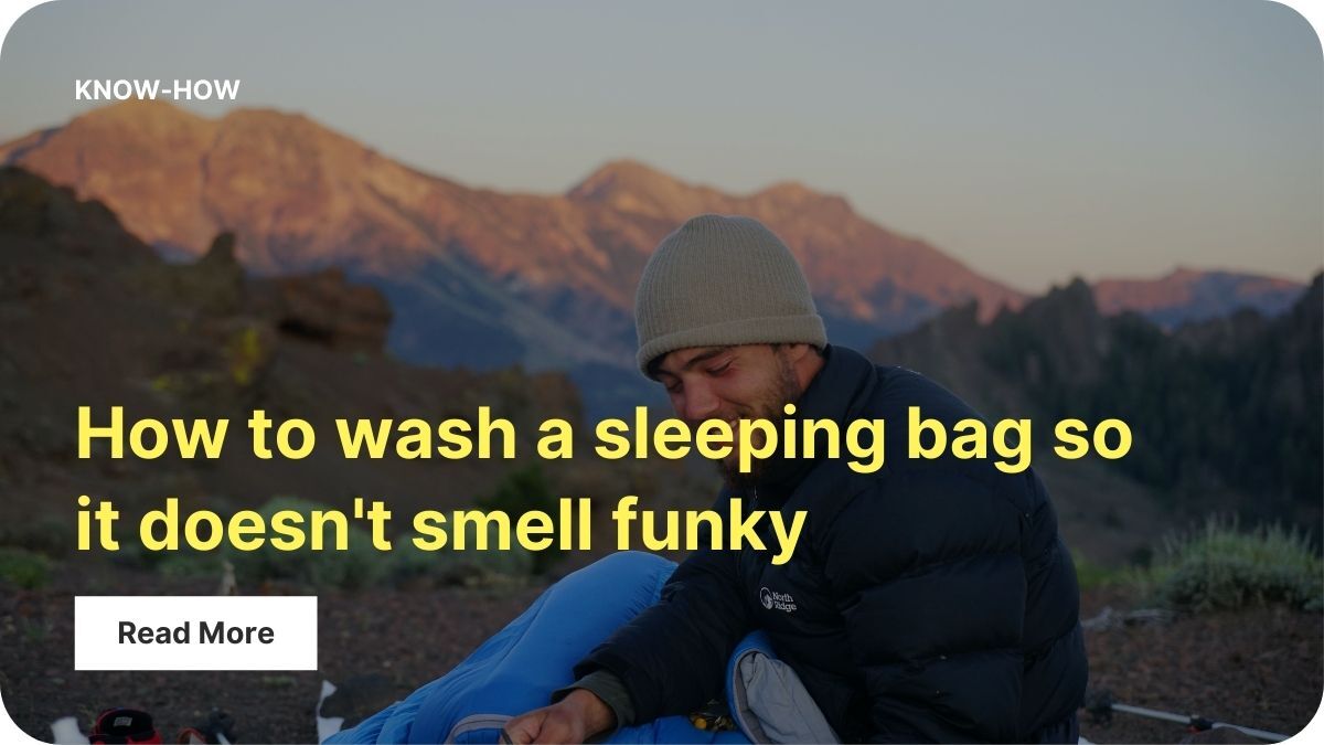 A person in a hat and fleece is half in a sleeping bag while using a camp stove. They're outside and there are mountains in the background.