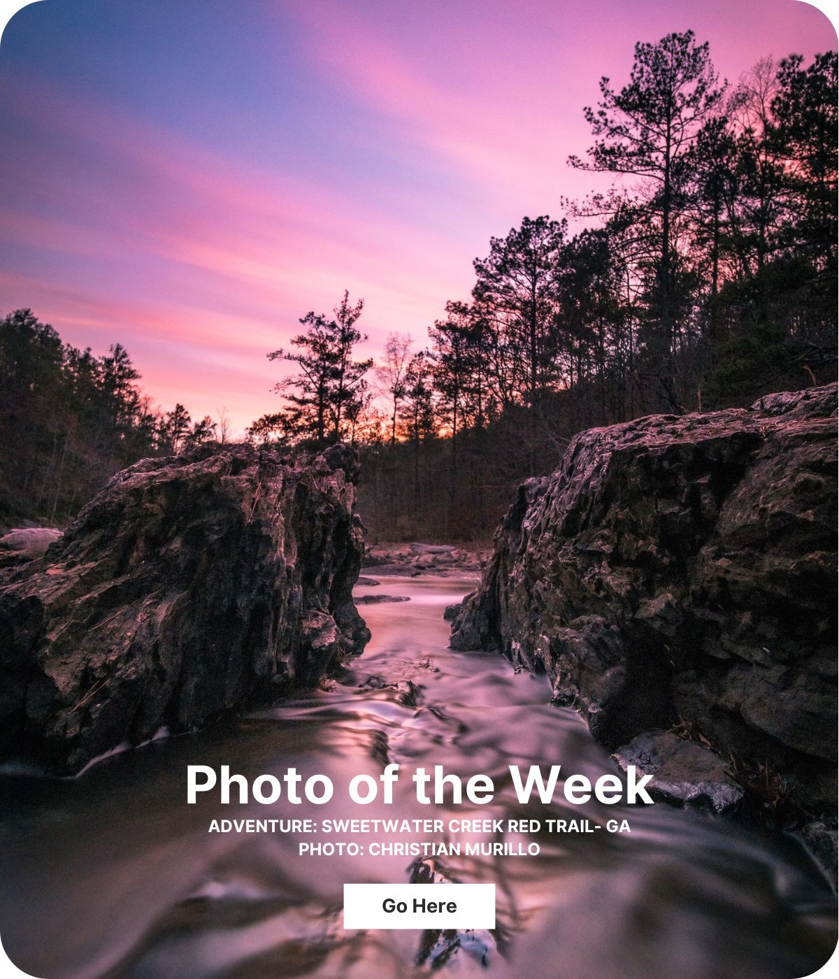 The camera is close to the water between two large rocks and the shoreline is lined with trees. The sky is blue and pink with whispy clouds.