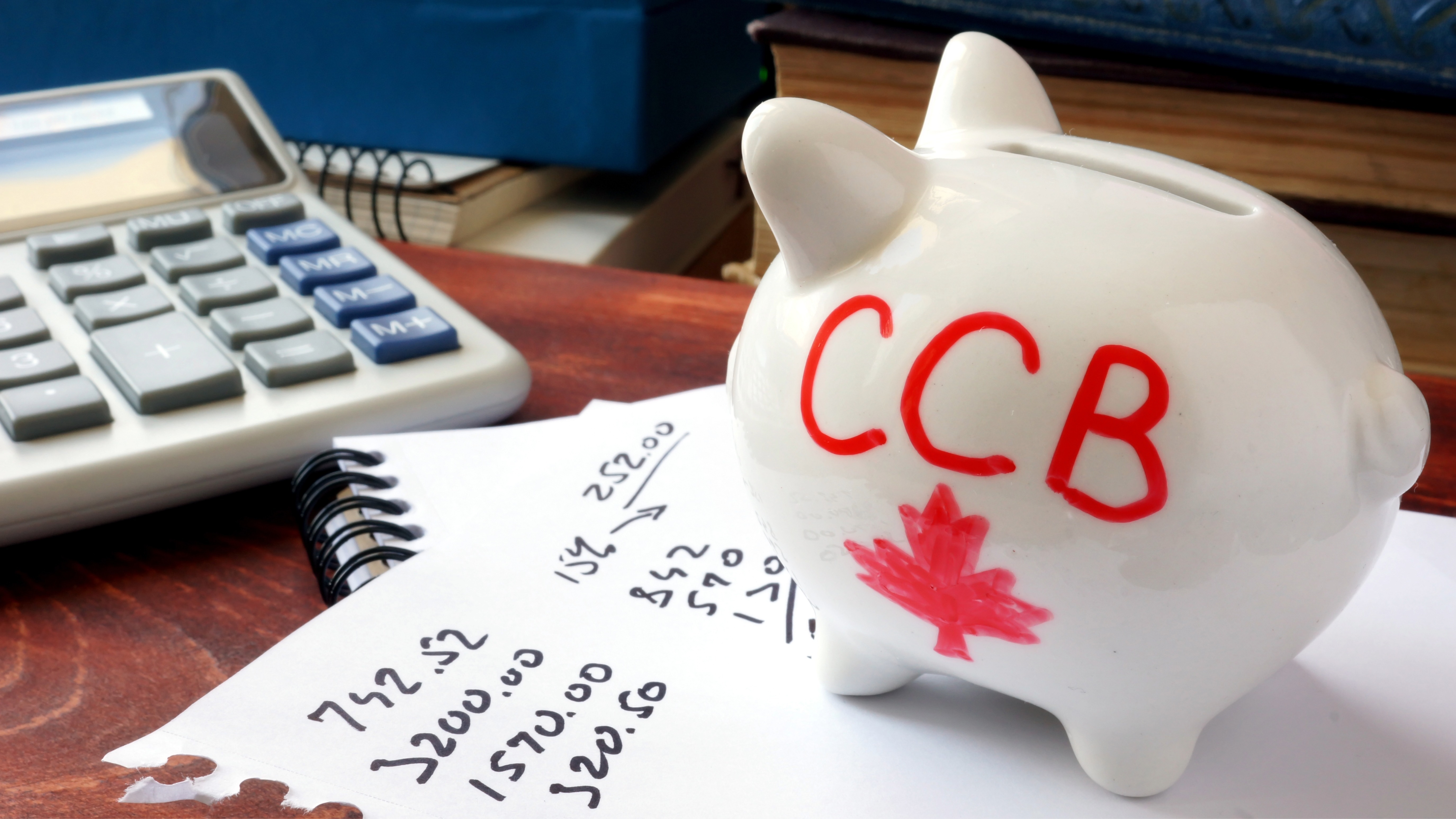 Desk with  a calculator, and notepad on it plus a white piggy bank with red writing that reads CCB and a maple leaf. 