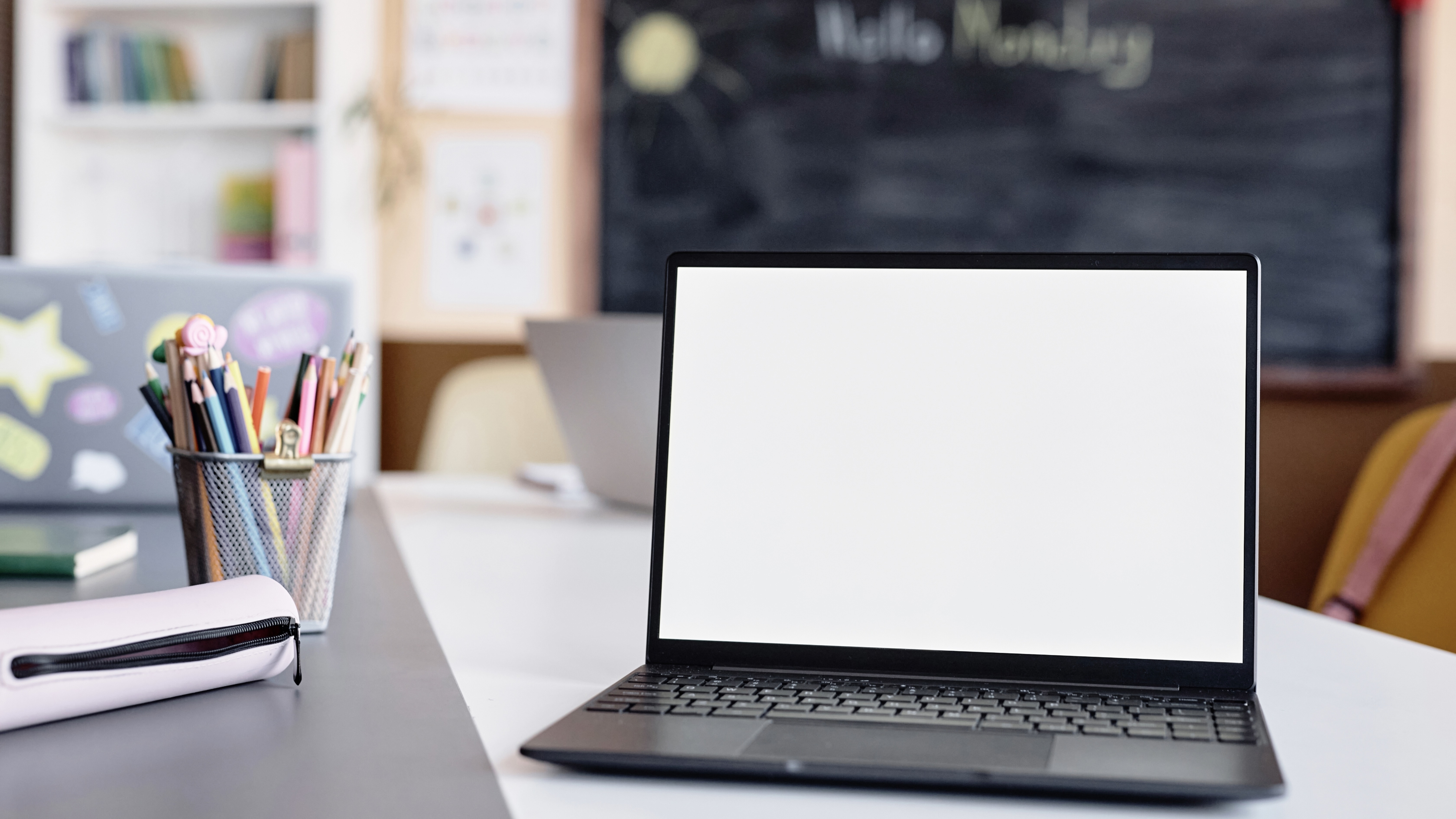 White desktop with a black open laptop on it. There is a white pencil case to the left as well as a metal pencil holder full of pens and pencils. 