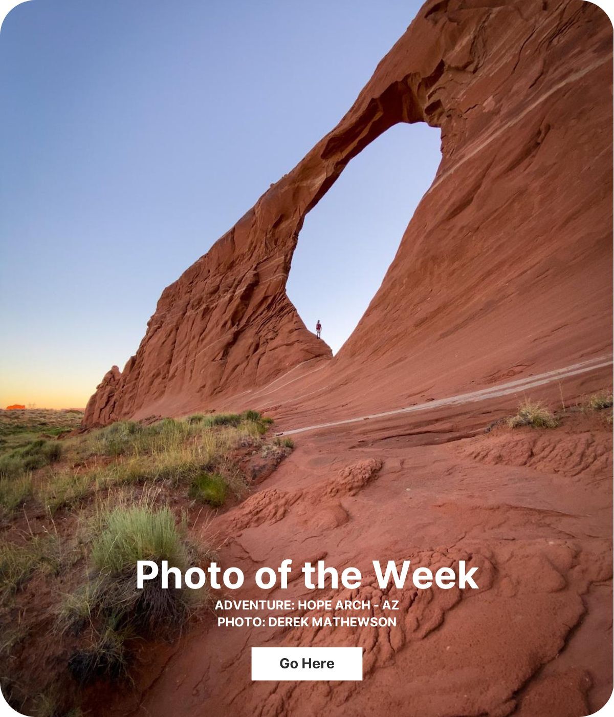 A tall red rock structure has an arch in the middle. A person stands under the arch. The sky is blue and turning yellow and orange at the horizon.