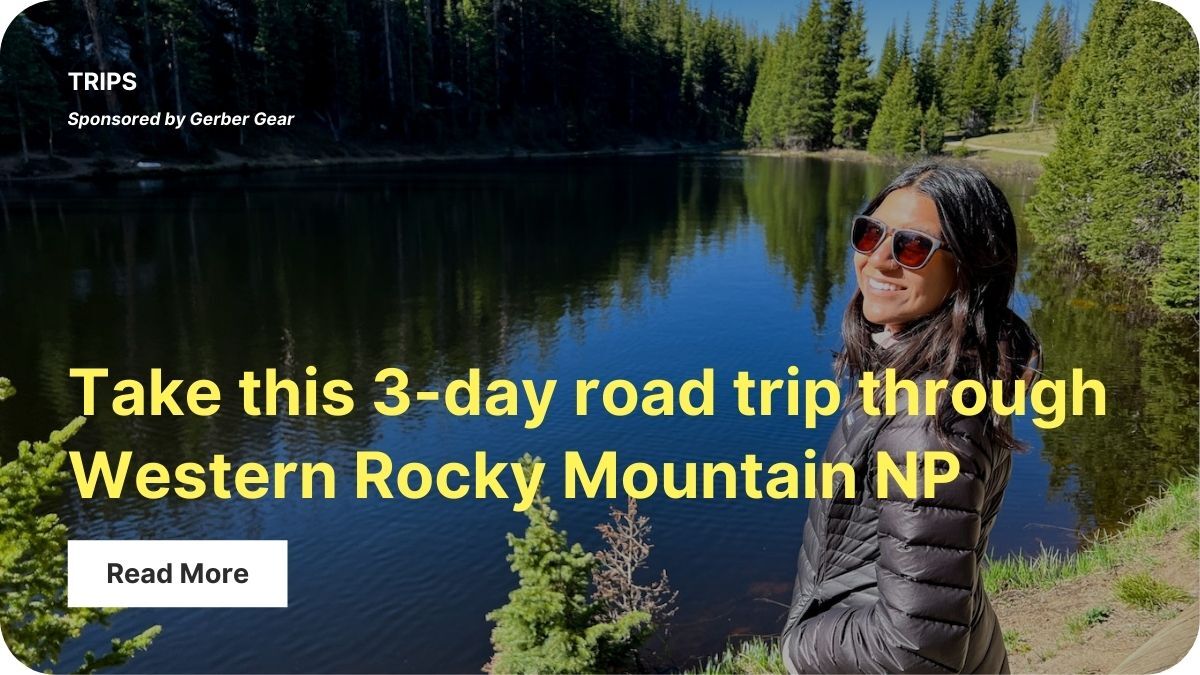 A person with long hair and sunglasses is wearing a black puffy jacket and standing next to a lake. 
