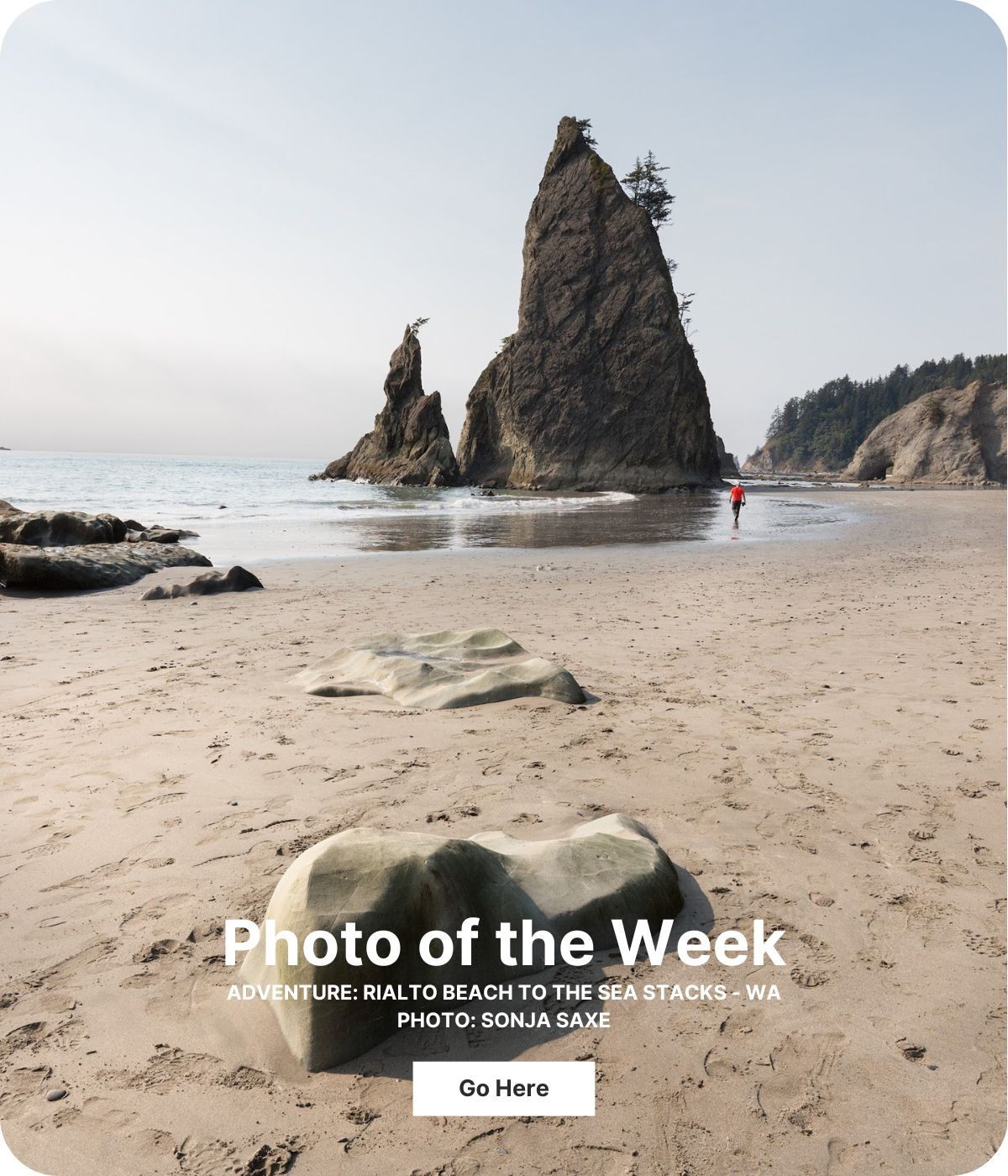 Two tall, triangular rocks rise out of water at a beach. A person in a red jacket is far away at the shoreline.