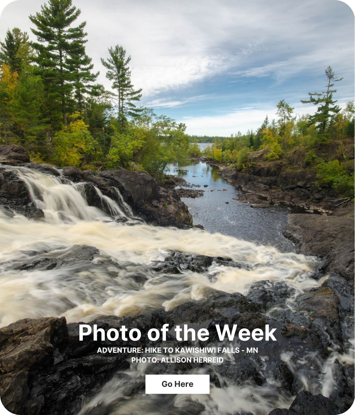 A waterfall cascades over dark rocks and flows into a river. Evergreen trees line the sides of the water.