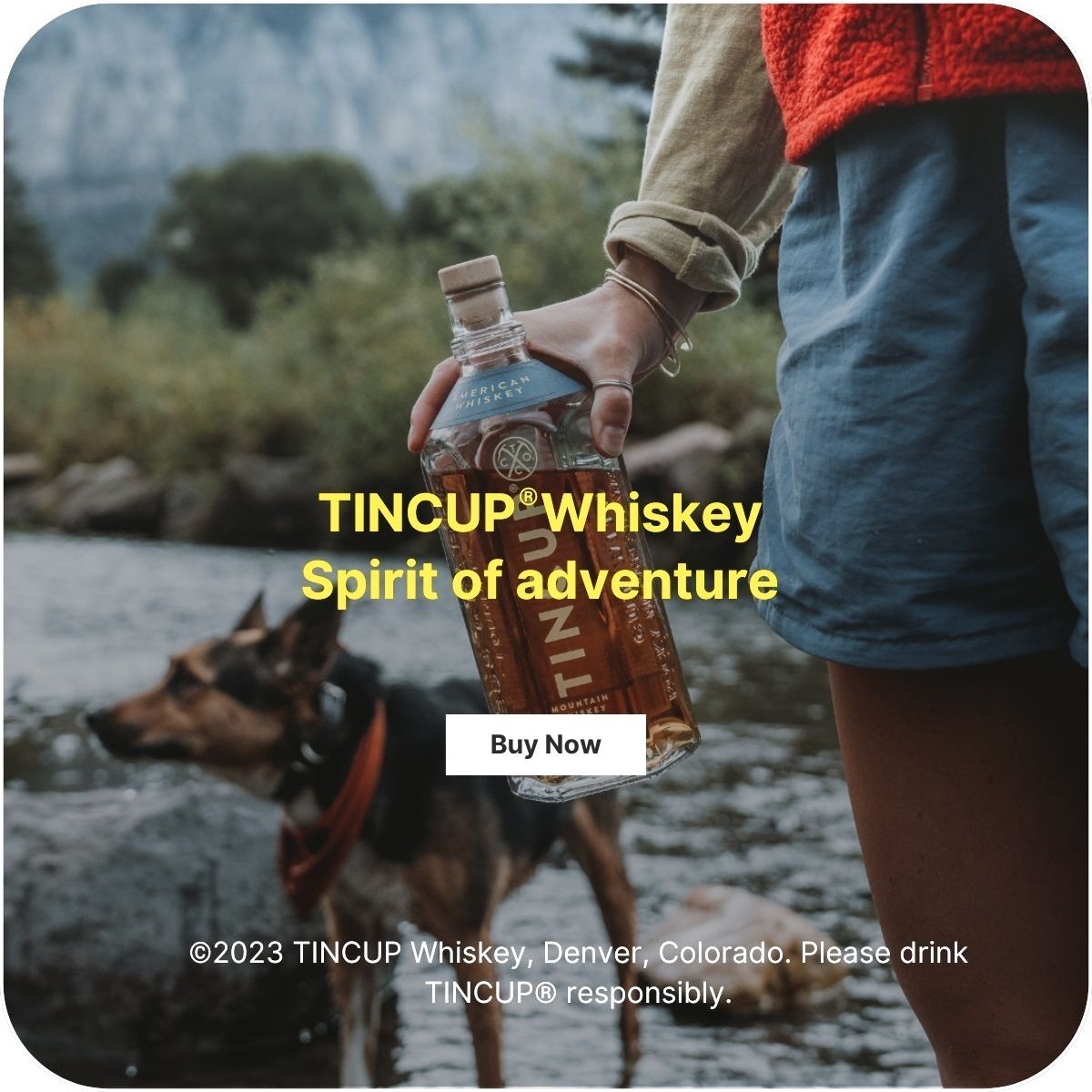 A close-up of a person holding a bottle of TINCUP Whiskey. They are standing next to a river and there is a dog out of focus behind the person.