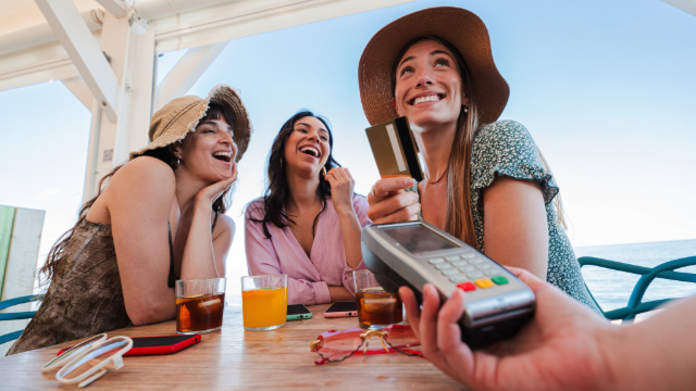 Three young women sitting at a table on a beach. One ha their credit card out and the server has the payment machine there. 