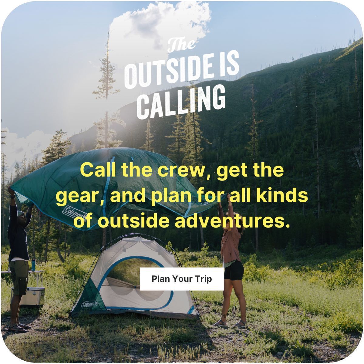 Two people are setting up a tent in a grassy area near a mountain. They're each holding onto one side of a rain tarp and have fluffed it up in the air to get it over the tent before installing it.