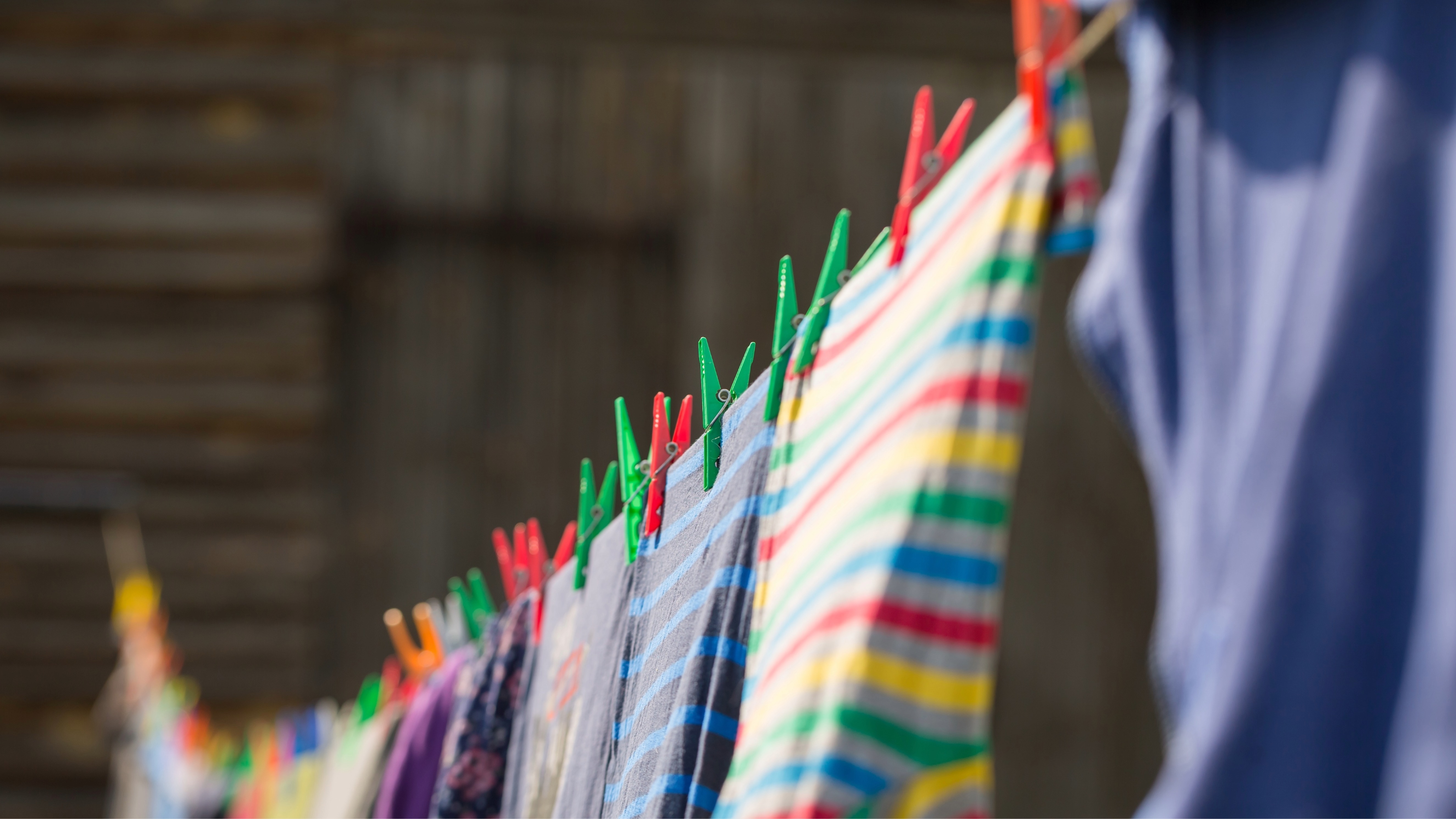 Multi-coloured clothes hanging on an outdoor clothesline. They are held on by red, green, orange, blue and yellow plastic clothes pins. 